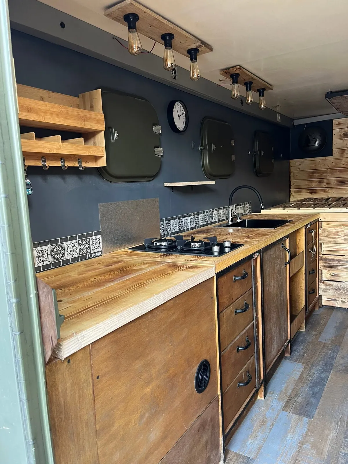 Intérieur du shelter : cuisine en bois, plaque gaz deux feux, évier noir et hublots