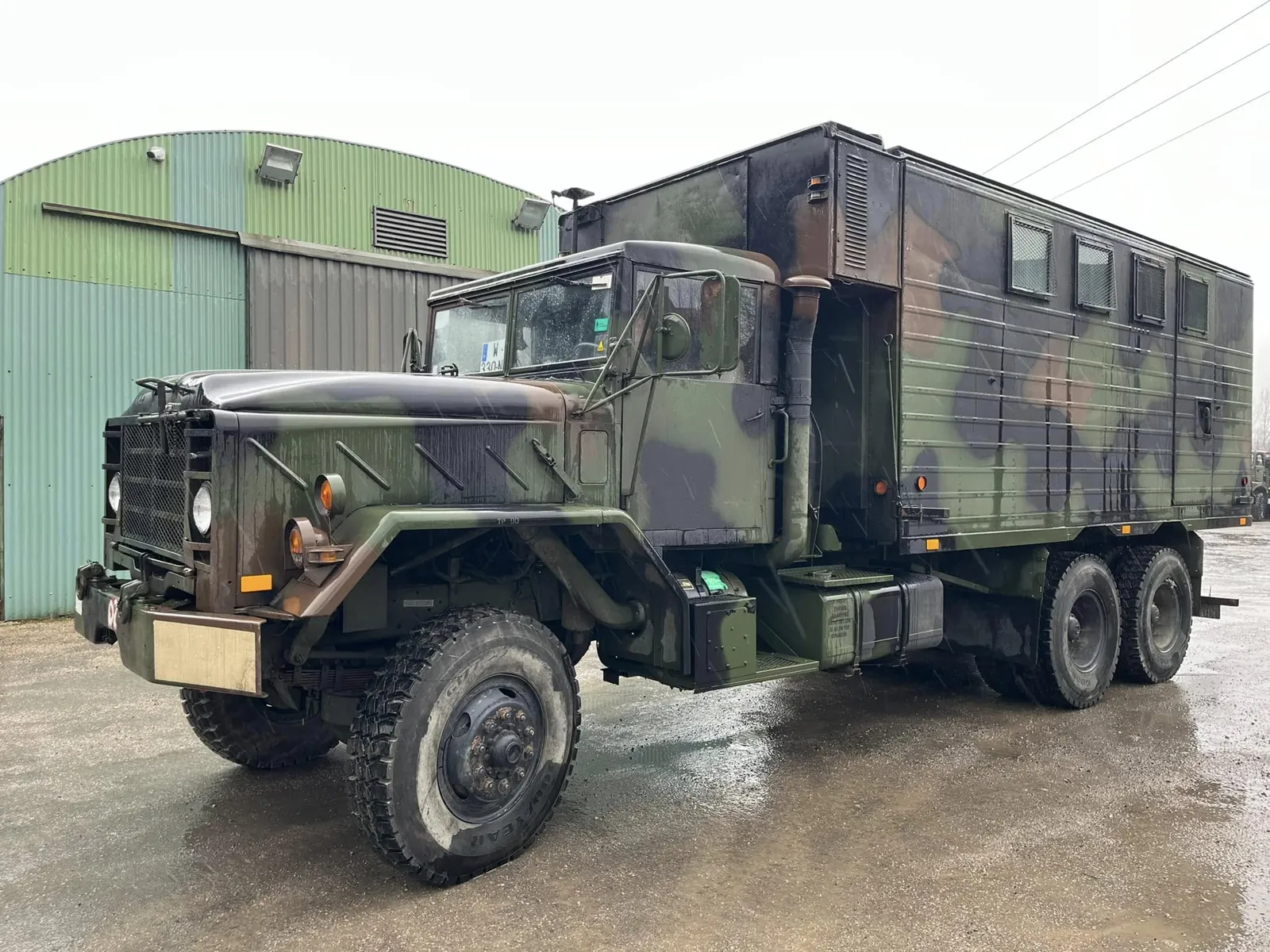 Camion AMG M934 à caisse fermée camouflée, trois quarts avant, devant un hangar vert