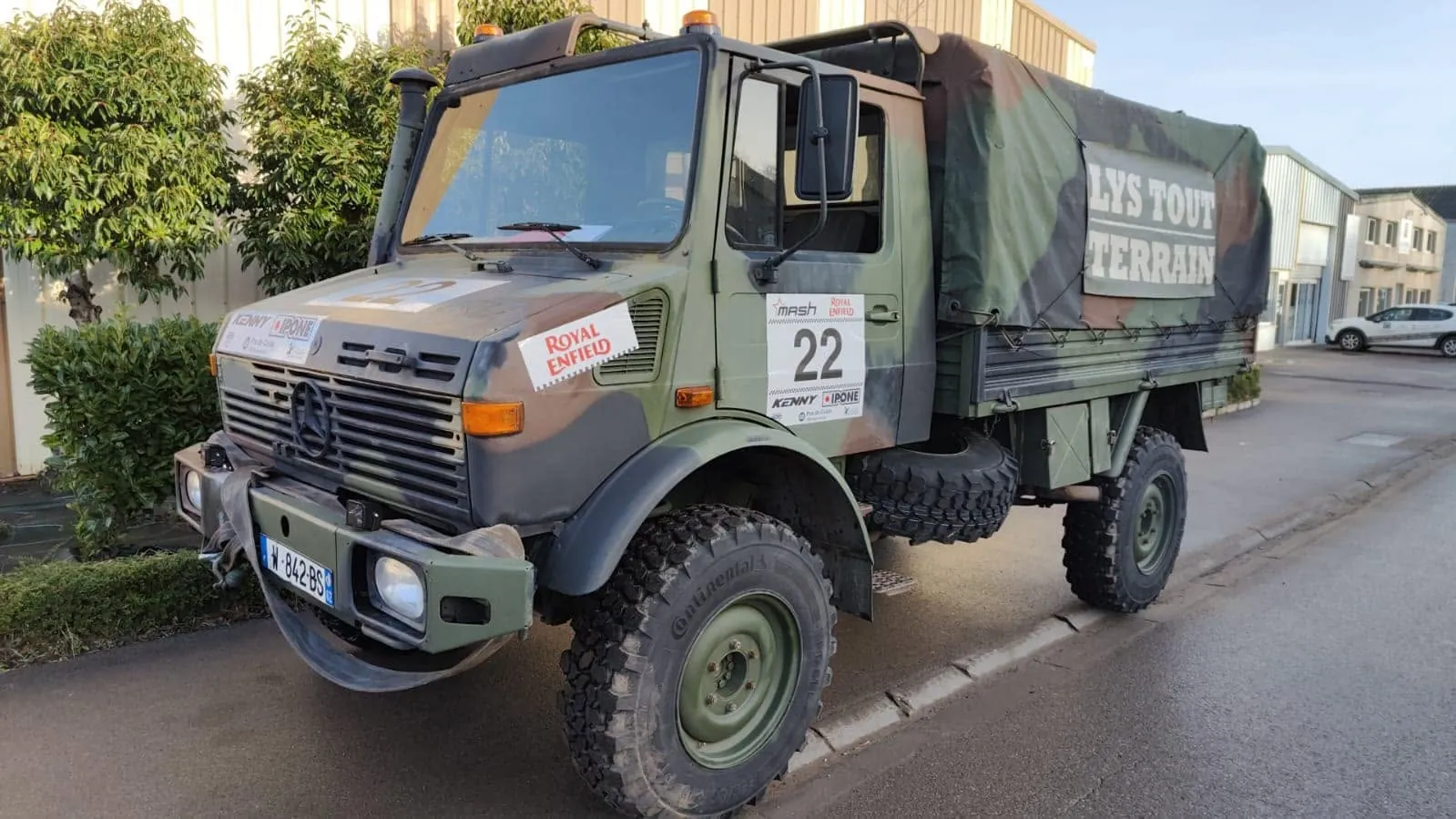 Mercedes Unimog camouflé numéro 22, bâche marquée Lys Tout Terrain, stationné le long d’un trottoir