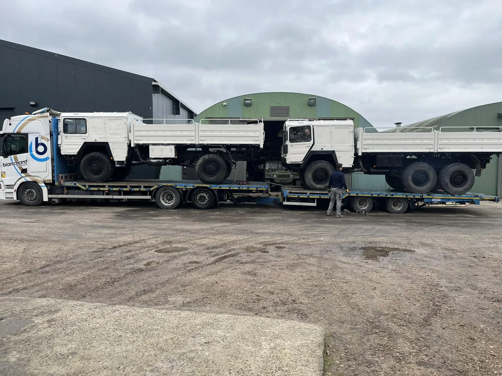 Deux camions MAN blancs à plateau ridelles sur une semi-remorque surbaissée devant les hangars