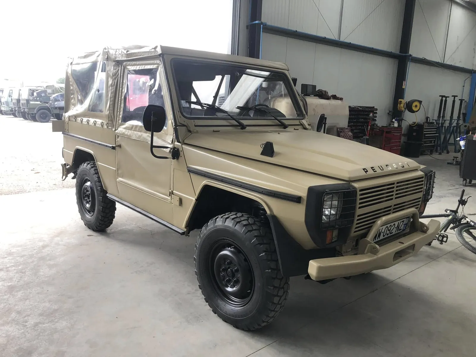 Peugeot P4 beige sable bâché, jantes noires, vu de trois quarts avant sous le hangar